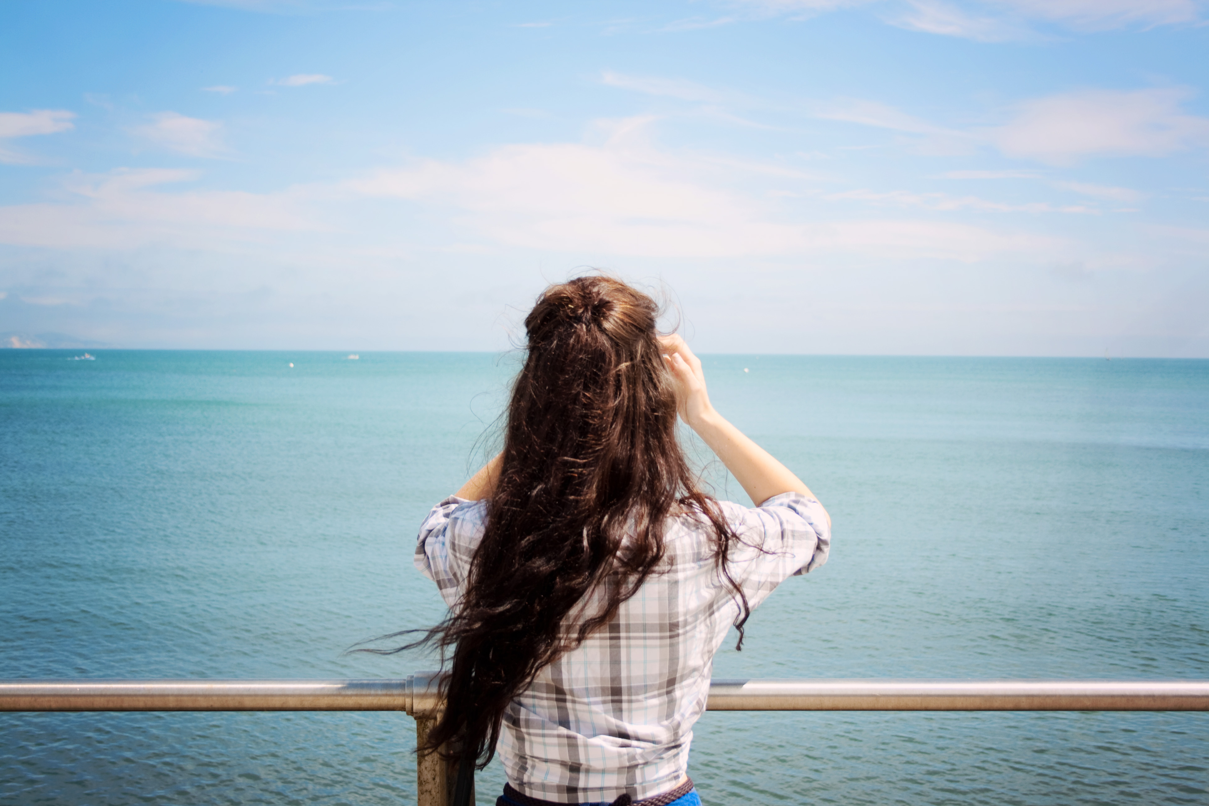 Teen girl with long brunette hair looking out to sea. Blue water and steel railings.