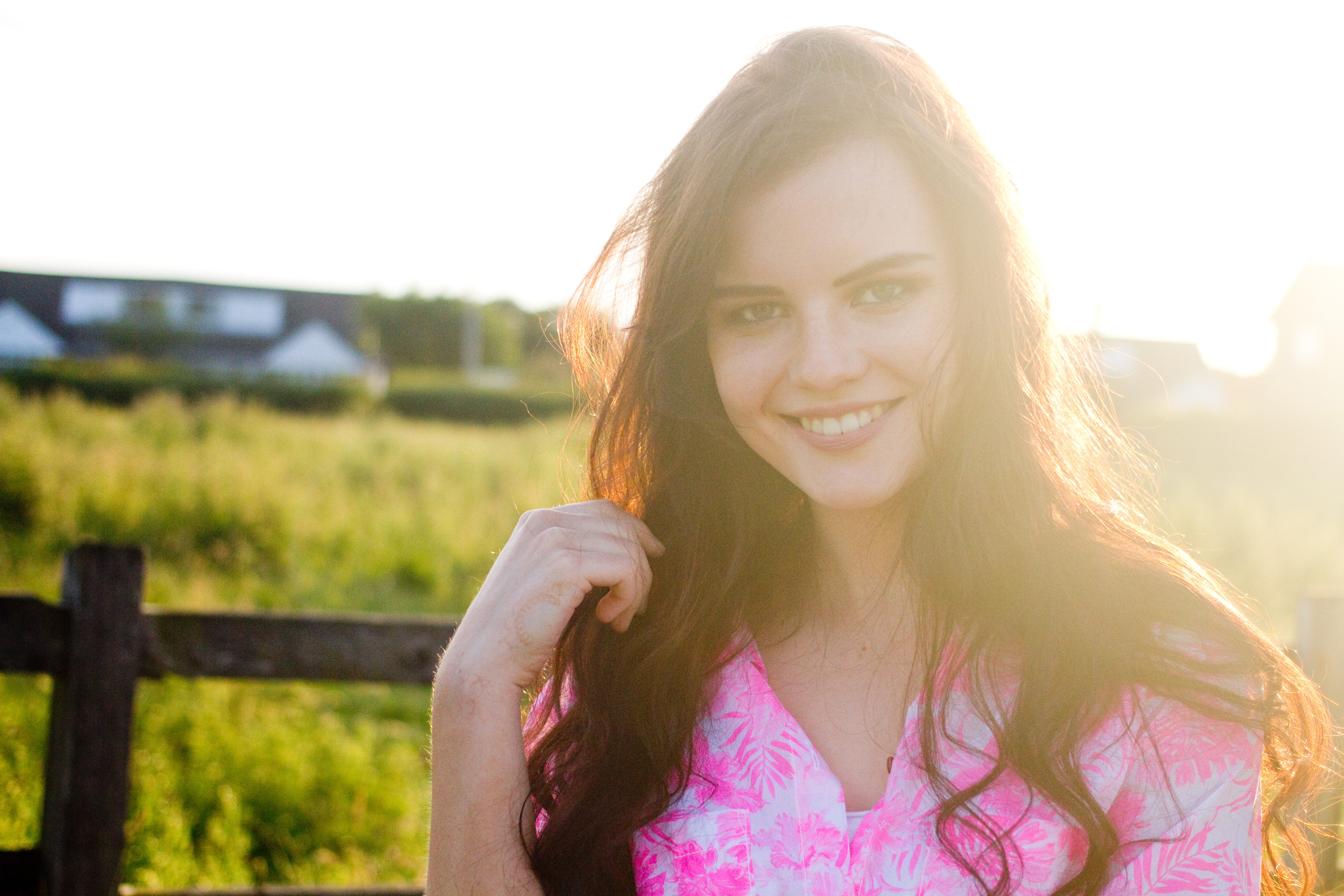 Backlit portrait of teen girl wearing bright pink patterned shirt. Green grass and fence in background.